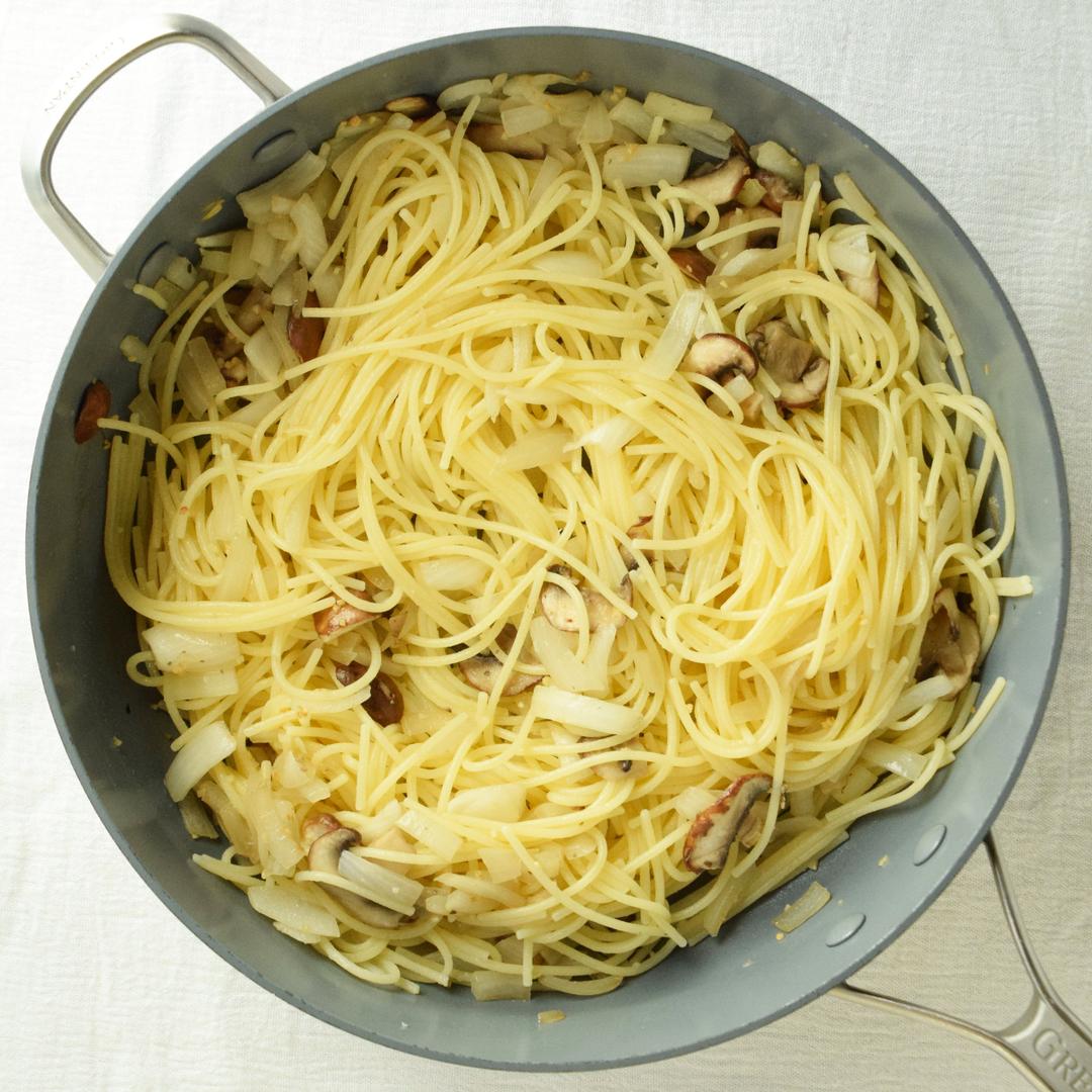 cooked spaghetti added to onions and mushrooms in saute pan